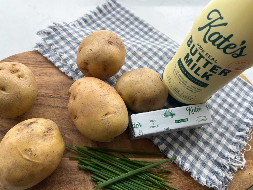 Potatoes sitting on a cutting board with a cloth, a stick of Kate's Butter, and Kate's Buttermilk