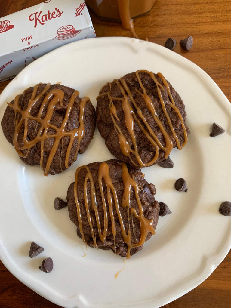 chocolate chip cookies with caramel drizzle on a plate with chocolate chips scattered around and a stick of kate's butter